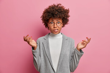 Photo of puzzled young African American woman entrepreneur with curly hair raises hands shrugs...