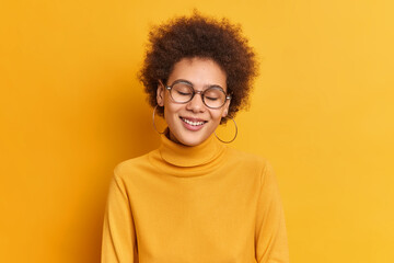 Fototapeta na wymiar Horizontal shot of happy sincere millennial girl with curly hair natural beauty smiles gently with closed eyes gets compliment enjoys life wears casual poloneck isolated over yellow background