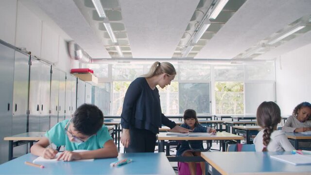 Female school teacher walking between desks, checking pupils doing task in classroom, giving help to diverse school kids. Dolly shot. Education or teaching concept