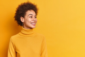 Pretty teenage girl with Afro hair looks gladfully aside smiles broadly being amused by someone dressed in casual turtleneck isolated over yellow background with blank empty space for your text