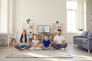 Lockdown pastime good for physical and mental health. Calm parents and children doing yoga at home. Happy family with kids sitting cross-legged on warm floor rug and practicing meditation together