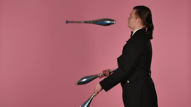 Side view of professional circus artist masterly juggles with clubs in the studio on a pink background. Man in black and white suit practices concentration, dexterity and skill. Close up. Slow motion.