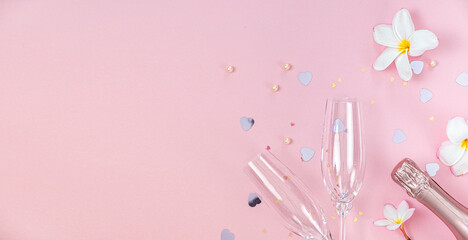 Two empty champagne glasses and bottle of champagne with white frangipani flowers and small heart decoration on pink background, copy space