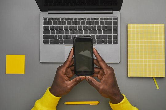 Graphic gray and yellow background of African-American male hands holding smartphone with blank screen over laptop while working at desk, top down view, copy space