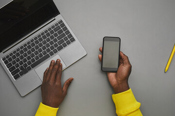 Minimal grey background of African-American male hands holding smartphone with blank screen over laptop while working at desk, top down view, copy space