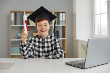 Happy smiling pupil in mortar board holding course completion certificate, sitting at desk with laptop computer. Portrait of smart child, successful student, online school graduate. Education concept