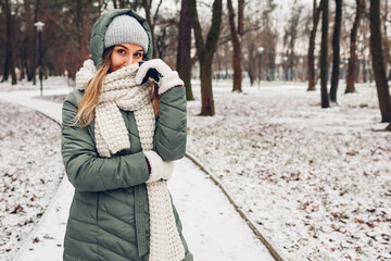 Winter fashion. Young woman wearing long green coat with scarf, hat, mittens in snowy park.