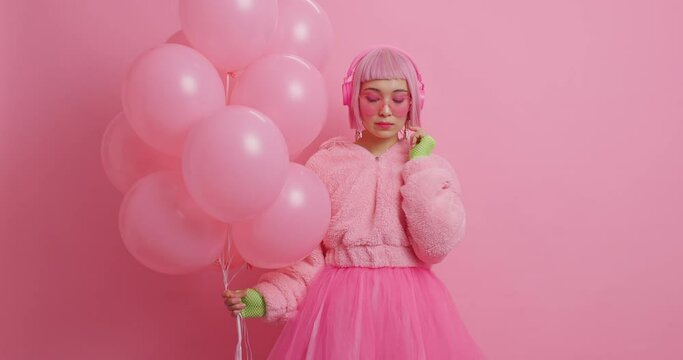 Sad lonely bored woman with bob pink hair looks down listens lyrics song via stereo headphones dressed in fashionable coat and skirt awaits for holiday holds inflated helium balloons poses indoor