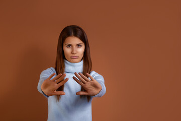 Displeased young european woman wearing light blue sweater shows stop sign prohibition symbol, keeps both palms forward to camera, pouts her lips, copy space on right, isolated on brown background.