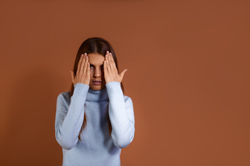 Scared young caucasian woman wearing light blue sweater covers face with hands, peeks out from under the palm, has mouth open, does not want to deal with something, isolated over brown background.