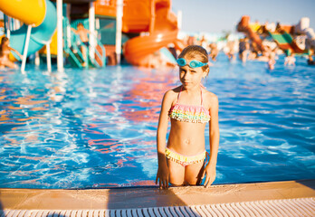 Funny girl in blue goggles for swimming stands in a pool with clean transparent water looking at the camera and enjoying the warm summer sun