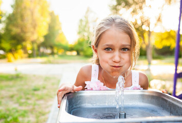 Cheerful wonderful girl drinks cool fresh water from a small fountain in a summer warm sunny park on a long-awaited vacation
