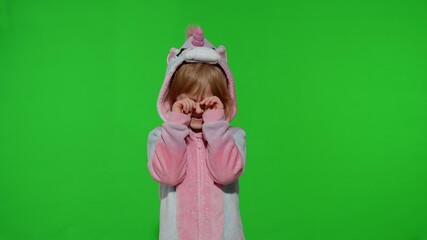 Young little blonde child 5-6 years old in unicorn costume crying, wiping tears with her hands on chroma key background. Portrait of kid girl animator in unicorn pajamas. Copy space