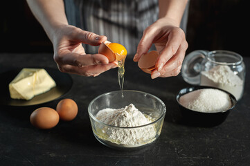 The process of preparing the dough. The woman breaks the egg and adds it to a bowl of flour. In women's hands an egg and a knife. Ingredients for the dough on a dark background.