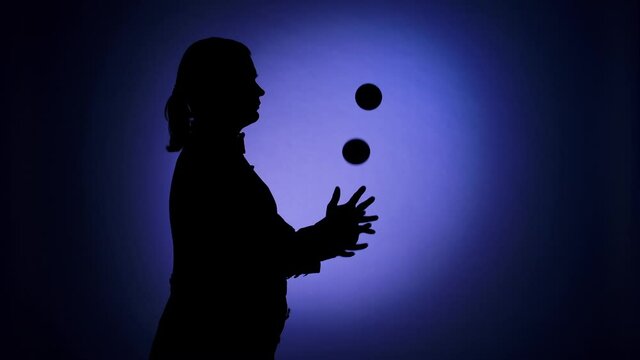 Side view black silhouette of a man juggling balls in the spotlight. Professional circus performer with an entertaining show program. Blue backdrop illuminated by studio light. Close up. Slow motion.