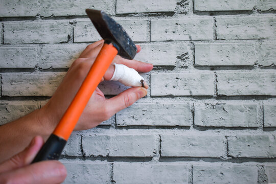 Hand With Bandaged Finger Drives Nail Into The Wall, Instead Of Nail It Hits Finger With Hammer. Man Drives Nail Into Wall, Bruises His Finger. Hand With Injured Finger Holds Hammer On Brick Wall Back