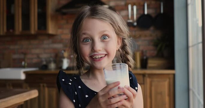 Head shot portrait cheerful little 7s girl standing alone in domestic kitchen holding glass drinks sweet yoghurt, milk mustache funny adorable face. Healthy lifestyle, beverage for kid growth concept