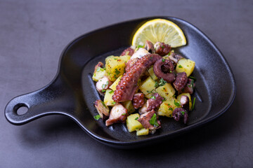 Warm salad with octopus, potatoes and lemon in a black small pan. Close-up, black background.