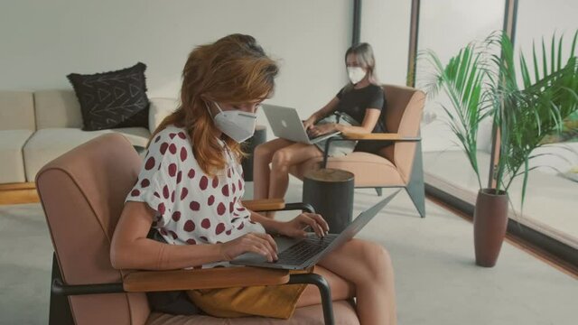 Two young female coworkers working in coworking space with modern interior maintaining social distancing while working on laptop wear N95 face masks to protect against the epidemic coronavirus