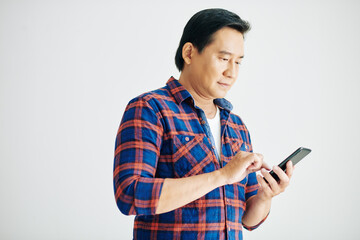 Smiling middle-aged man in plaid shirt answering text messages on smartphone or checking mobile...