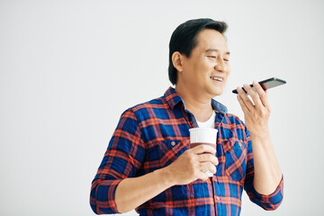 Smiling Asian man in plaid shirt drinking take out coffee and recording voice message