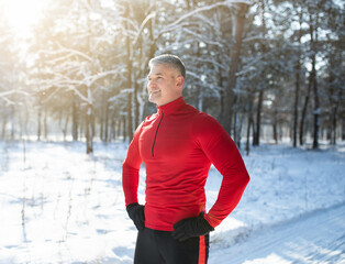 Happy senior man in warm sports clothes taking break after jogging at snowy winter forest. Seasonal outside activities
