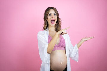 Young beautiful brunette woman pregnant expecting baby over isolated pink background surprised, showing and pointing something that is on her hand