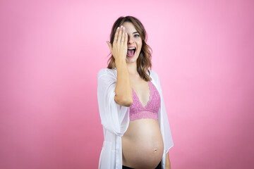 Obraz premium Young beautiful brunette woman pregnant expecting baby over isolated pink background covering one eye with hand, confident smile on face and surprise emotion.