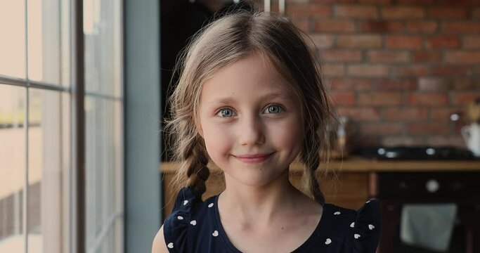 Head shot cute small 7s girl standing in kitchen smiles looks at camera. Adorable caucasian preschool child with pigtail poses indoors. Happy childhood, new Alpha generation offspring portrait concept