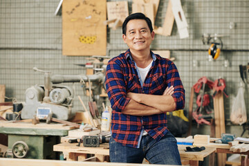 Cheerful confident carpenter in plaid shirt standing in his carpentry workshop with arms folded
