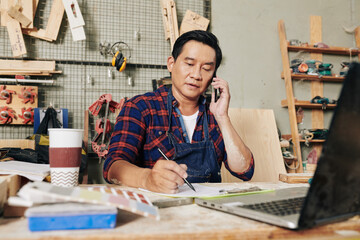 Portrait of mature serious carpenter sitting at desk in his workshop, talking on phone with customer and taking notes in planner