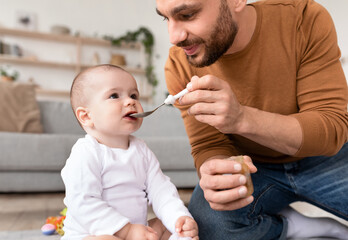 Loving Young Father Spoon Feeding His Little Baby Sitting Indoors