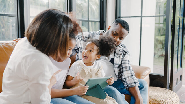 Young African American family and daughter happy using tablet at home. Mom, Dad, and child relax with little girl watching movie on sofa in living room. Funny parent and lovely child are having fun.