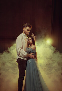 Fantasy couple hugging in dark room full white smoke. Image of lovers king and queen. Medieval male prince in golden crown, vintage costume clothing. Girl Princess in long glamorous blue dress gown