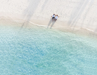 Young couple traveler sitting and relaxing at beautiful tropical white sand beach with wave foam and transparent sea, Summer vacation and Travel background Top view from drone