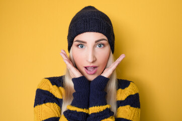 Shocked beautiful woman standing with positive surprised face expressing, model wearing woolen cap and sweater, isolated on yellow background. Crazy pretty girl