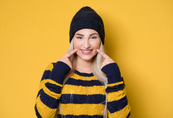 Smile young woman shows her cheeks and teeth, model wearing woolen cap and sweater, isolated on yellow background. Smiling pretty girl gesturing