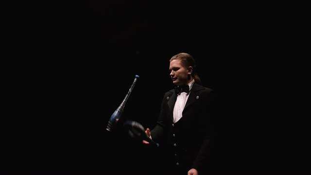 Camera rotates around circus juggler using pins to juggle. A man in a black suit performs exciting tricks in a dark backlit studio. Orbital shot close up. Slow motion.