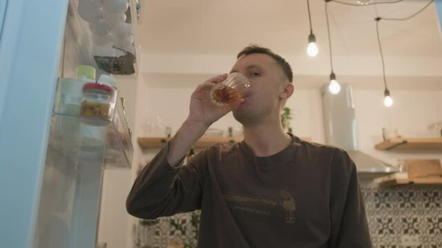 A young man takes a glass of beer from the refrigerator at home, drinks a couple of sips and puts the glass back, closes the refrigerator. Medium close-up