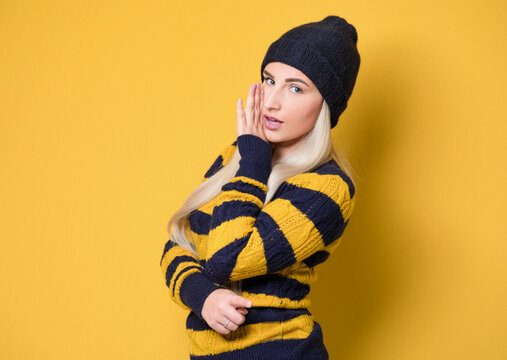 Woman holding hand near mouth and telling secret, model wearing woolen cap and sweater, isolated on yellow background. Gossip concept