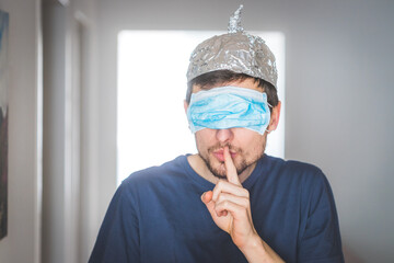 Conspiracy theory concept: Young man with face mask over the eyes and aluminum hat is making a...