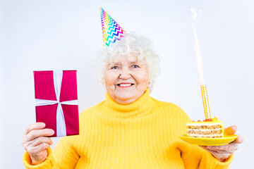 older woman with a gift wear yellow sweater and horn cap on a white background holding plate with cake with fireworks
