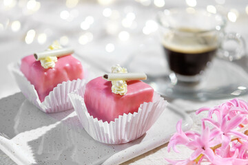 Valentine petit fours with marzipan icing and cream flowers. Espresso coffee in glass cup. Garland of lights on ivory, off white textile tablecloth. Fragrant pink hyacinth flower. Happy Valentine's