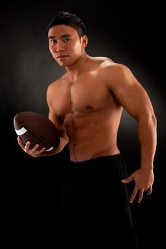 Muscular american football player holding ball against dark background.