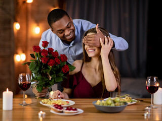 Romantic Surprise. Happy black man covering girlfriend's eyes and giving roses bouquet