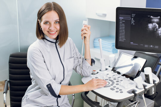Close up portrait of professional smiling young woman doctor sonographer sitting near modern ultrasound scanner machine and holding transducer