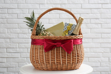 Fototapeta na wymiar Wicker gift basket with pineapple and bottle of champagne on table near white brick wall