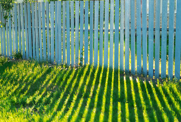The shadow of the old wooden blue fence is reflected on the green grass on a sunny summer day. A long, linear, perspective shadow of a picket fence on the lawn, in the garden.