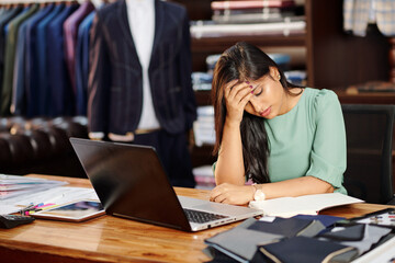 Stressed or tired young female atelier owner sitting at table with opened laptop and waiting for orders