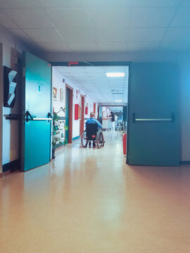 clinic with a senior in wheelchair. At the beginning of the corridor there is a loney patient, on the background some nurses are working using a crash cart (vertical image with copy space)